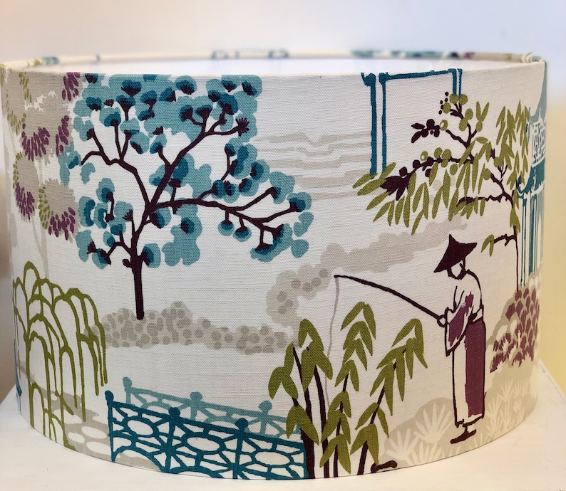 May include: A white lampshade with a blue, green, and purple floral pattern. The pattern features trees, a bridge, and a person fishing.