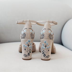 May include: A pair of beige block-heel shoes with ankle straps, featuring delicate embroidery of light blue flowers and dark green stems on the back of the heels and the rear upper section of the shoes, showcasing a clean and structured design.