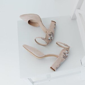 May include: A pair of beige suede block-heel pumps featuring a pointed toe, an adjustable ankle strap with a metal buckle, and delicate floral embroidery in light blue and black thread detailing on the heel and back of the shoe.