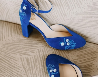 May include: A pair of blue suede heels with floral embroidery. The heels have a block heel and an ankle strap.