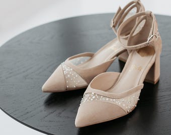 May include: A pair of beige suede block-heel shoes featuring a pointed toe, an ankle strap with a silver-tone buckle, and sheer mesh side panels embellished with small white beads and sequins that create a delicate, shimmering texture throughout.