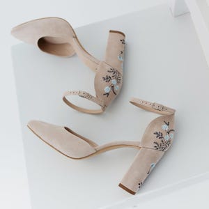 May include: A pair of beige suede block-heel pumps featuring a pointed toe, an adjustable ankle strap with a metal buckle, and delicate floral embroidery in shades of light blue and dark green on the heel and back of the shoe.