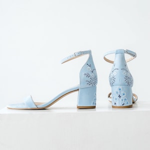 Op de afbeelding: Een paar lichtblauwe sandalen met bloemenborduursel. De sandalen hebben een blokhak en een enkelband.