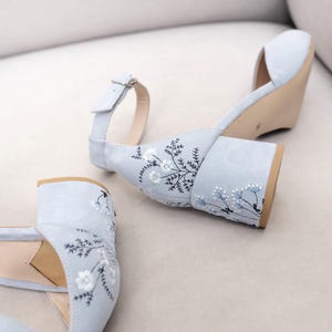 Puede incluir: Un par de sandalias de tac&oacute;n de ante azul claro con bordados florales. Las sandalias tienen la punta cerrada y una correa de tobillo con hebilla.