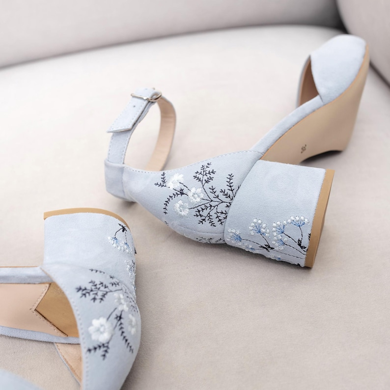 Puede incluir: Un par de sandalias de tac&oacute;n de ante azul claro con bordados florales. Las sandalias tienen la punta cerrada y una correa de tobillo con hebilla.