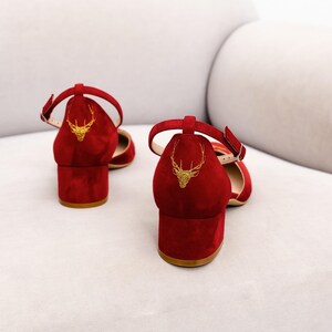 K&ouml;nnte beinhalten: Ein Paar rote Wildleder-Schuhe mit Blockabsatz und Kn&ouml;chelriemen. Jeder Absatz ist auf der R&uuml;ckseite mit einem goldenen, gestickten Hirschkopf-Motiv versehen. Die Schuhe haben eine geschlossene Zehenpartie, einen niedrigen Blockabsatz und einen Riemen.
