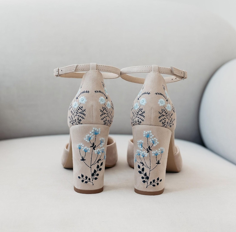 May include: A pair of beige block-heel shoes with ankle straps, featuring delicate embroidery of light blue flowers and dark green stems on the back of the heels and the rear upper section of the shoes, showcasing a clean and structured design.
