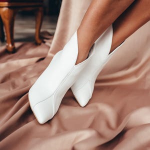 May include: A pair of white ankle boots featuring a pointed toe and a distinct V-shaped cutout at the front opening. The boots have a smooth finish and a structured silhouette that extends upwards, showcasing clean stitching lines along the sides.