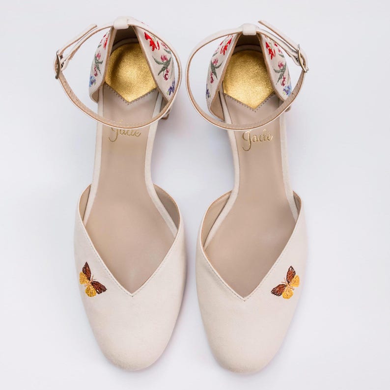 May include: A pair of cream-colored pointed-toe shoes with ankle straps. The shoes feature embroidered butterflies on the toes and floral embroidery on the heel counters. The insoles are gold with the brand name Joie printed in gold script lettering.