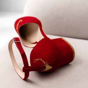K&ouml;nnte beinhalten: Ein tiefroter Wildlederschuh mit Blockabsatz und Kn&ouml;chelriemen mit silberfarbener Schnalle. Die Au&szlig;enseite des Schuhs ist mit goldfarbenen Stickereien verziert, die den Umriss eines Hirschgeweihs an der Ferse und im Zehenbereich darstellen.