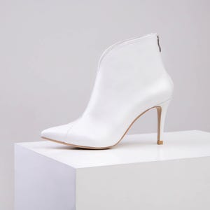May include: A white ankle boot with a pointed toe and a high stiletto heel. The boot features a deep V-shaped cutout along the front shaft and a vertical seam running down the centre. A small silver zip is visible at the top of the back heel.
