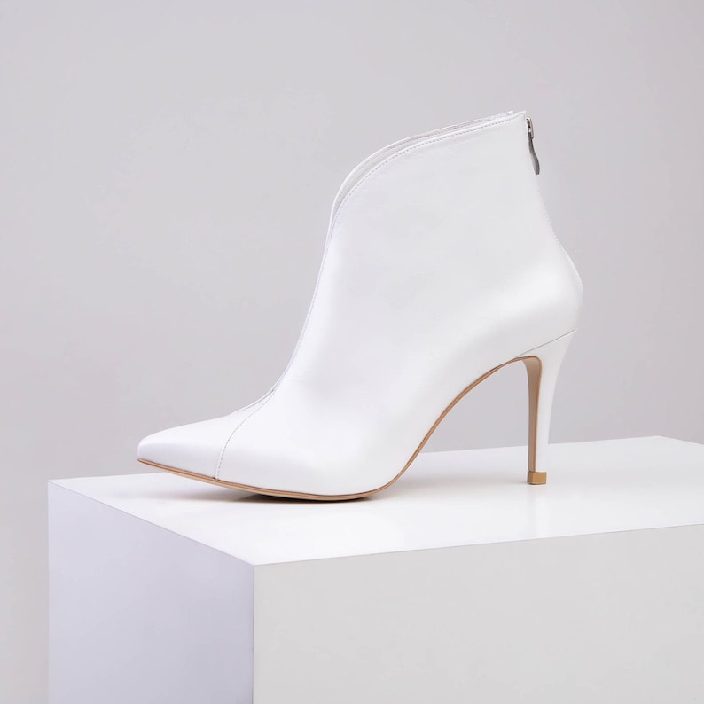 May include: A white ankle boot with a pointed toe and a high stiletto heel. The boot features a deep V-shaped cutout along the front shaft and a vertical seam running down the centre. A small silver zip is visible at the top of the back heel.
