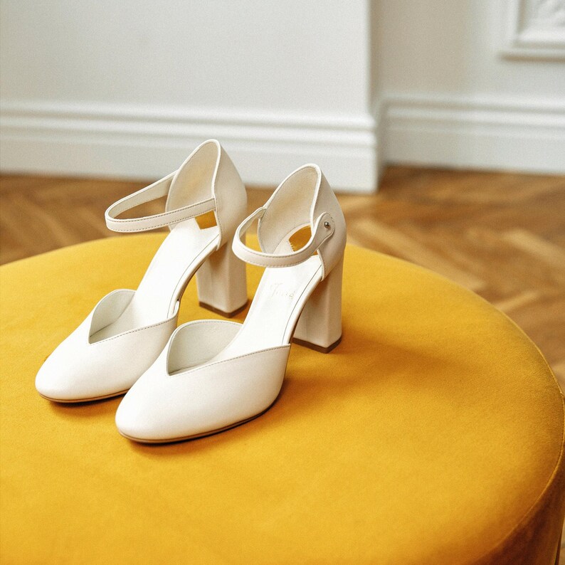 May include: A pair of off-white closed-toe pumps featuring a block heel, a rounded toe box with a V-shaped cutout at the vamp, and a thin ankle strap secured with a small metal buckle. The shoes have a smooth finish and a structured heel design.