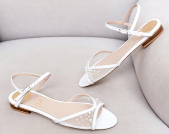 May include: A pair of white flat sandals featuring a sheer mesh toe strap adorned with small white pearls. The sandals have a thin ankle strap with a silver-toned metal buckle, a low heel, and a light-coloured insole with the brand name Jojo printed.