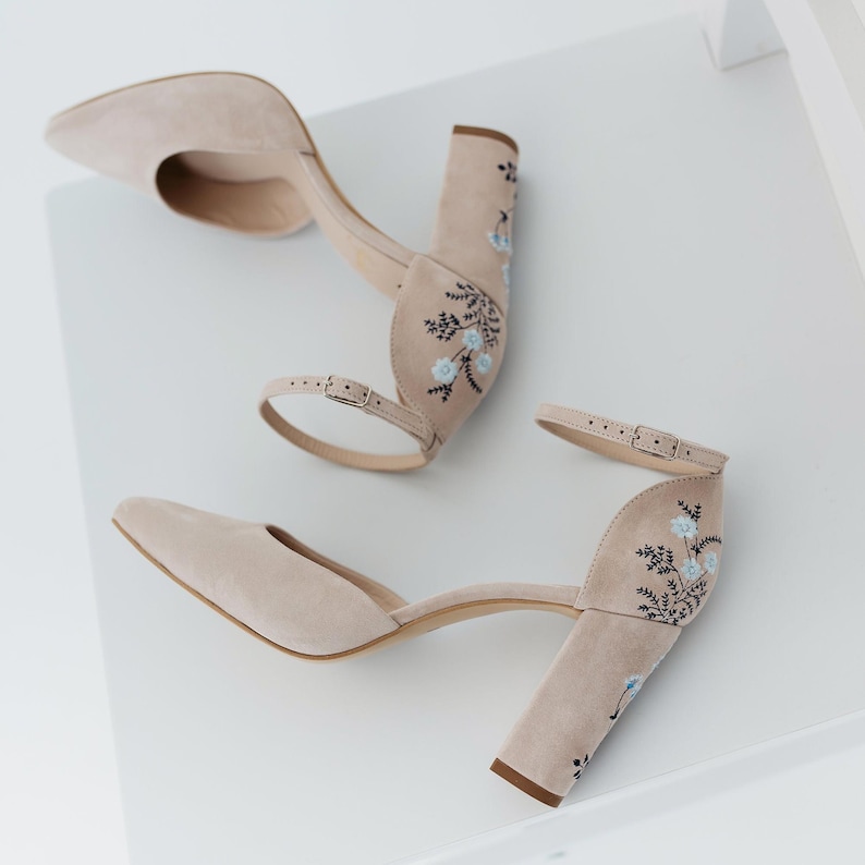 May include: A pair of beige suede block-heel pumps featuring a pointed toe, an adjustable ankle strap with a metal buckle, and delicate floral embroidery in shades of light blue and dark green on the heel and back of the shoe.