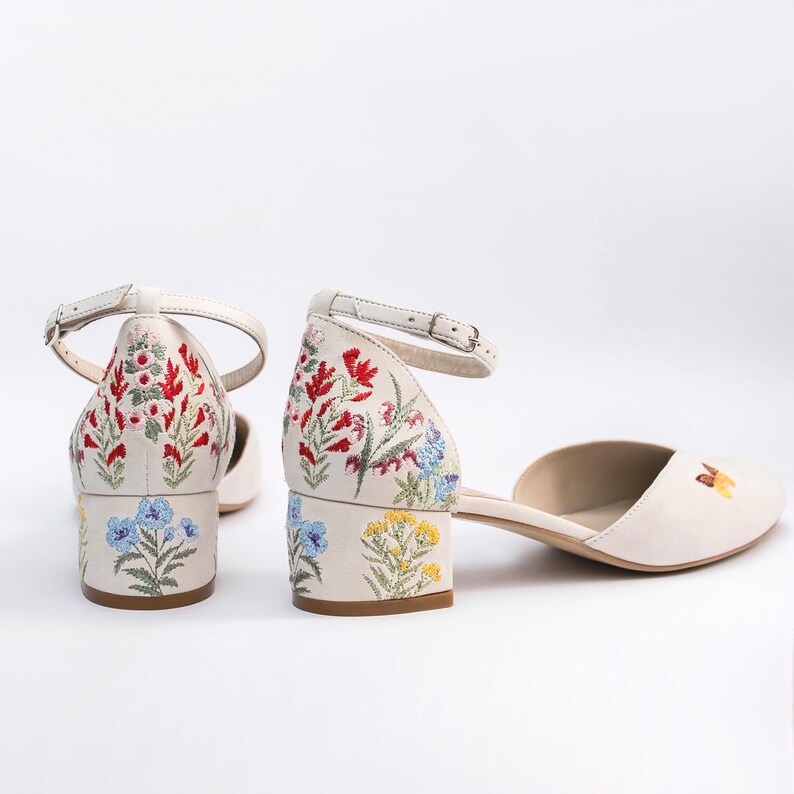 May include: A pair of off-white block-heeled shoes featuring an ankle strap with a metal buckle. The heel and back of the shoes are decorated with colorful floral embroidery in red, blue, yellow, and green, with a small butterfly on the toe.