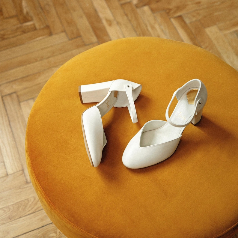 May include: A pair of white leather shoes with a rounded toe, a block heel, and a thin ankle strap. The shoes feature a cutout design on the sides and a gold-toned interior lining. The construction includes a secure buckle closure on the strap.