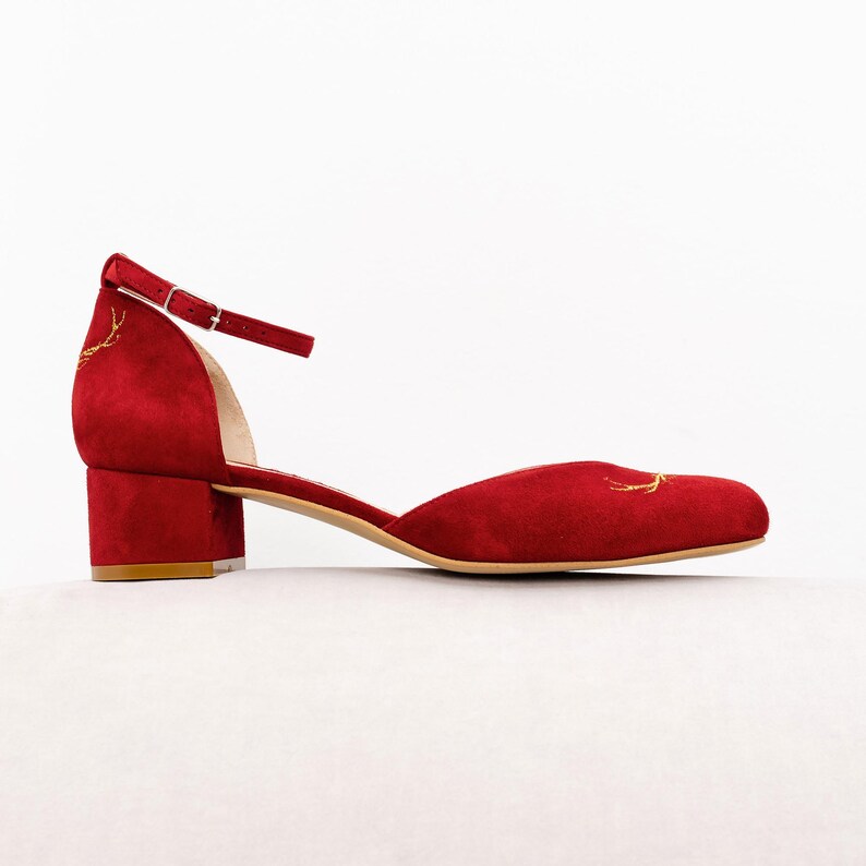 K&ouml;nnte beinhalten: Ein roter Wildleder-Schuh mit Kn&ouml;chelriemen und niedrigem Blockabsatz. Der Schuh hat eine abgerundete Zehenpartie und goldene Stickereien in Form von Hirschgeweihen an der Ferse und der Schuhspitze. Er besitzt einen d&uuml;nnen, verstellbaren Riemen.