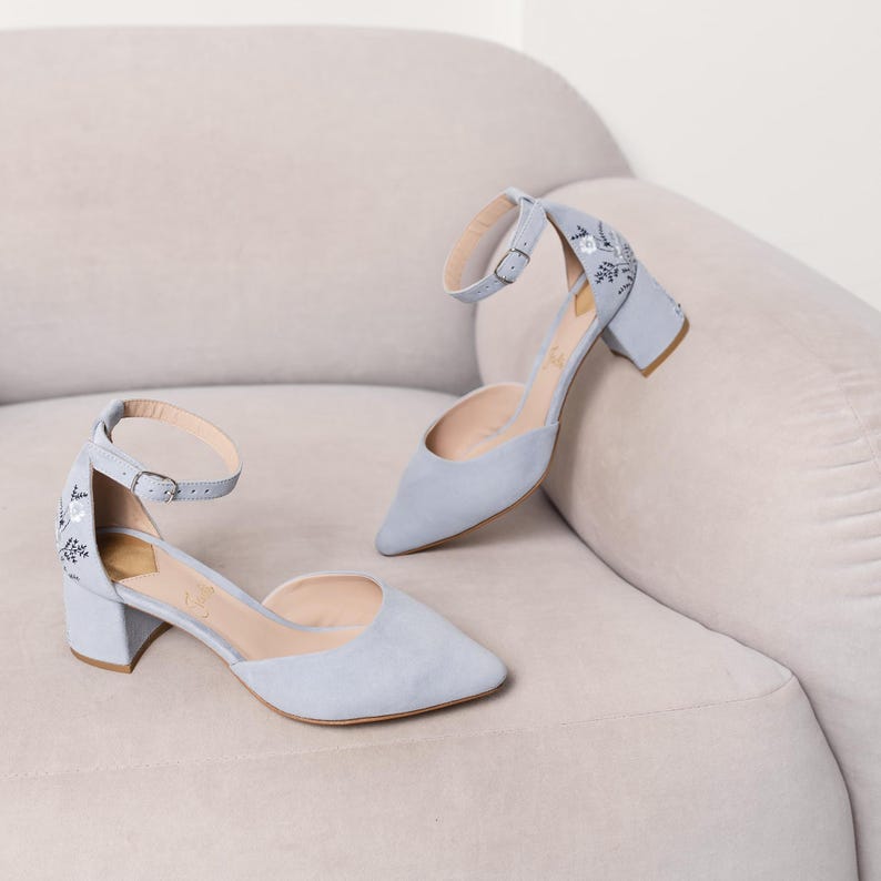 Puede incluir: Un par de zapatos de tac&oacute;n de ante azul claro con correas de tobillo y delicado bordado floral. Los zapatos tienen un tac&oacute;n de bloque y una punta puntiaguda.