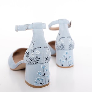 Puede incluir: Un par de zapatos de tac&oacute;n de ante azul claro con correas de tobillo. Los zapatos tienen un dise&ntilde;o de bordado floral en la parte posterior del tac&oacute;n.