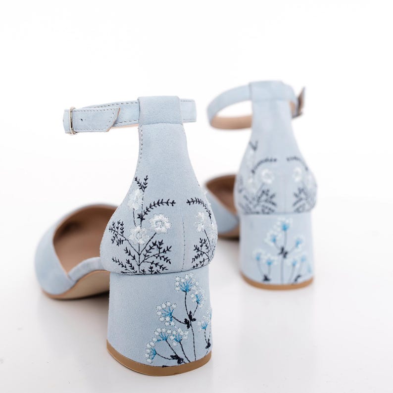 Puede incluir: Un par de zapatos de tac&oacute;n de ante azul claro con correas de tobillo. Los zapatos tienen un dise&ntilde;o de bordado floral en la parte posterior del tac&oacute;n.
