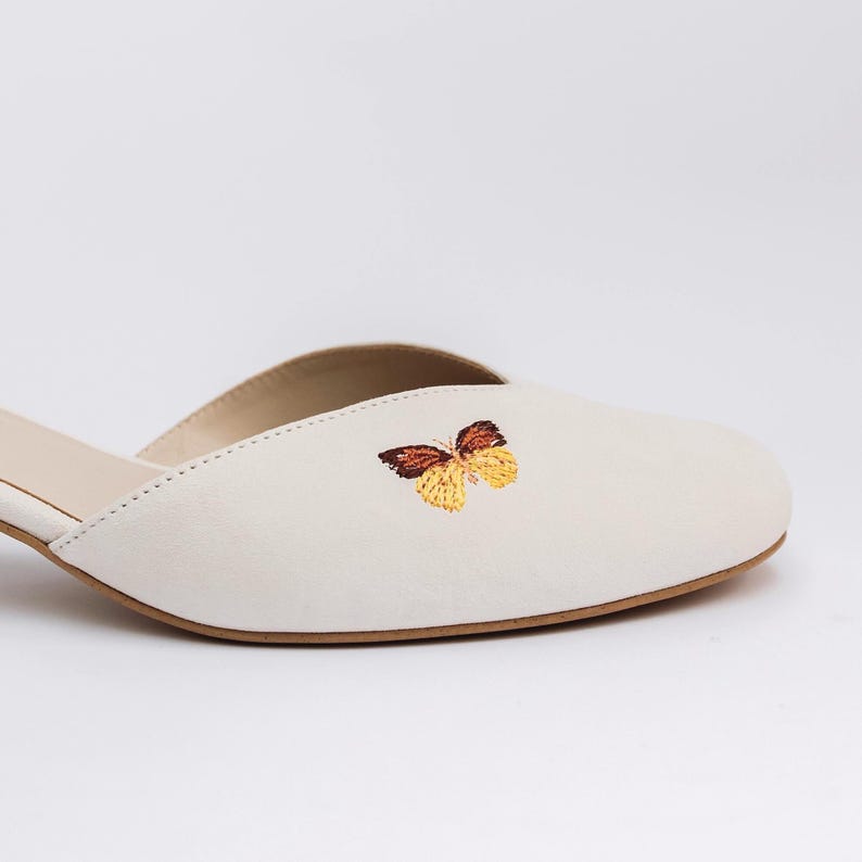 May include: A cream-colored slip-on mule shoe featuring a small, embroidered butterfly design on the upper. The butterfly has brown and yellow wings. The shoe has a rounded toe, a low-cut opening, and a thin, light brown sole edge.