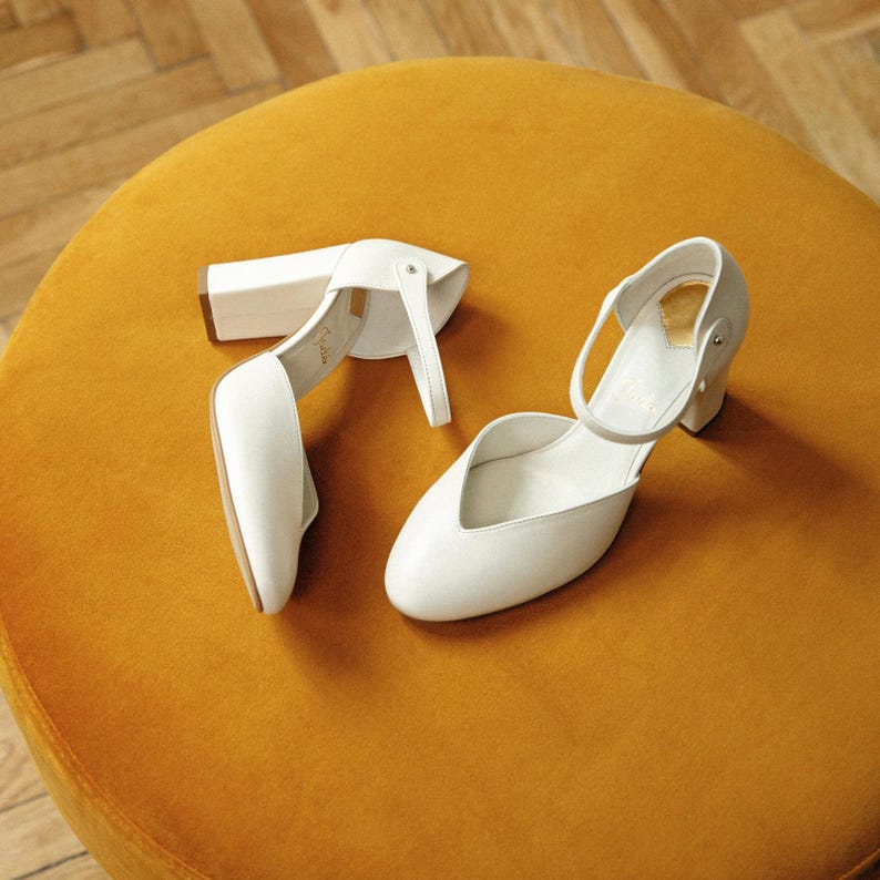 May include: A pair of white block-heeled shoes featuring a closed rounded toe, a low-cut vamp, and a thin ankle strap with a side buckle closure. The shoes have a smooth finish and a sturdy heel, providing a clean and structured appearance.