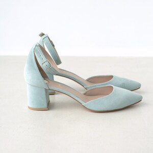 Puede incluir: Un par de zapatos de tac&oacute;n tipo d'Orsay en ante azul claro con punta afilada, tac&oacute;n bajo de bloque y correa ajustable al tobillo con hebilla de metal plateado. El forro interior es de color beige claro y el exterior tiene un acabado mate suave.