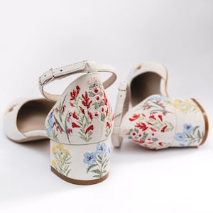 May include: A pair of off-white block-heeled shoes featuring an ankle strap with a metal buckle. The exterior of the shoes is decorated with colorful floral embroidery, showcasing red, pink, yellow, and blue flowers with green stems and leaves.