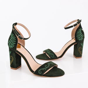 May include: A pair of dark green suede heeled sandals with an ankle strap. The sandals feature embroidered green leaves and vines on the straps and heels.