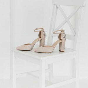 May include: A pair of beige suede high-heeled shoes with pointed toes and ankle straps. The heels feature delicate floral embroidery in shades of blue and green. The shoes are positioned on a white wooden chair with a cross-back design.