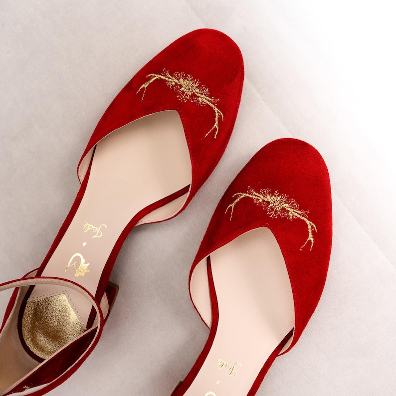 K&ouml;nnte beinhalten: Ein Paar rote Wildlederschuhe mit abgerundeter Spitze und einem goldfarbenen, gestickten Geweihmotiv auf der Vorderseite. Das Innere verf&uuml;gt &uuml;ber eine cremefarbene Innensohle mit dem goldenen Schriftzug Jade und einem kleinen goldenen Logo.