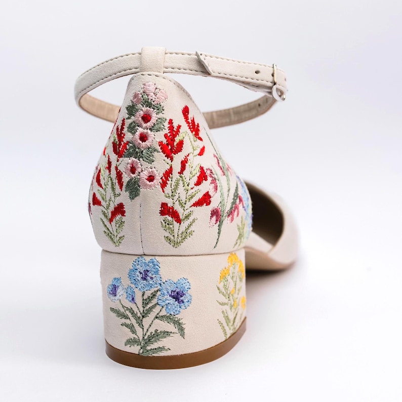 May include: An off-white block heel shoe featuring an ankle strap and intricate floral embroidery. The heel and back of the shoe are decorated with colorful stitched flowers in shades of red, pink, blue, and yellow with green stems and leaves.