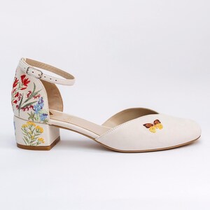 May include: An off-white ankle strap shoe with a low block heel. The heel and back feature colorful floral embroidery in red, blue, and yellow. A small, detailed butterfly is embroidered on the side of the toe box. The shoe has a metal buckle.
