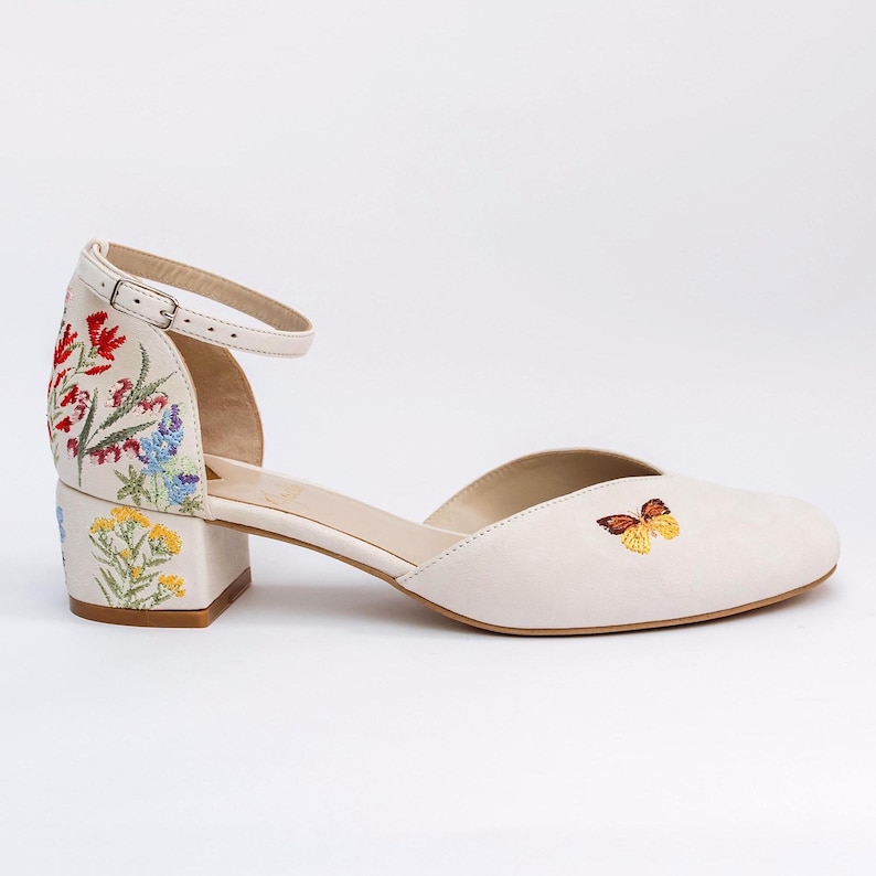 May include: An off-white ankle strap shoe with a low block heel. The heel and back feature colorful floral embroidery in red, blue, and yellow. A small, detailed butterfly is embroidered on the side of the toe box. The shoe has a metal buckle.