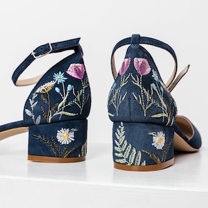 Könnte beinhalten: Ein Paar dunkelblaue Wildledersandalen mit Knöchelriemen. Die Absätze sind Blockabsätze und die Sandalen sind mit gestickten Blumenmustern in den Farben Rosa, Blau, Grün und Gelb verziert.