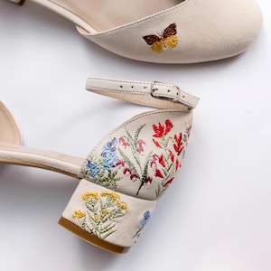 May include: A pair of cream-colored block heel shoes featuring an ankle strap with a metal buckle. The shoes are decorated with colorful floral embroidery on the heel and sides, and a small brown and yellow butterfly embroidered on the toe.