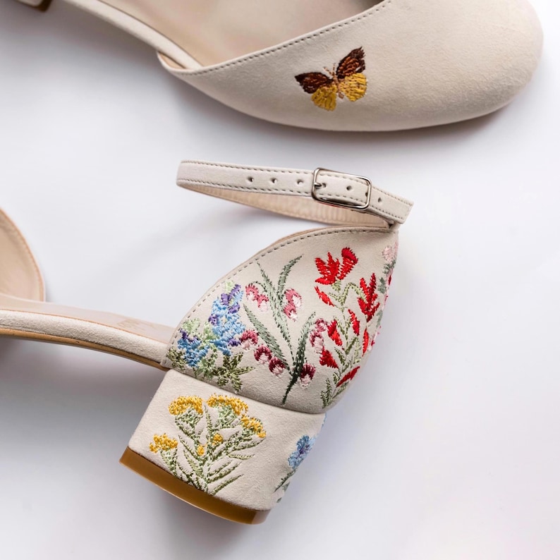 May include: A pair of cream-colored block heel shoes featuring an ankle strap with a metal buckle. The shoes are decorated with colorful floral embroidery on the heel and sides, and a small brown and yellow butterfly embroidered on the toe.