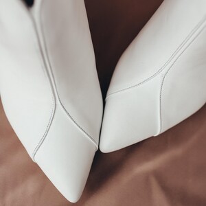 May include: A pair of white pointed-toe ankle boots featuring a smooth matte finish and a prominent vertical seam running down the centre of the vamp. The boots have a clean, structured design with visible tonal stitching along the seams.