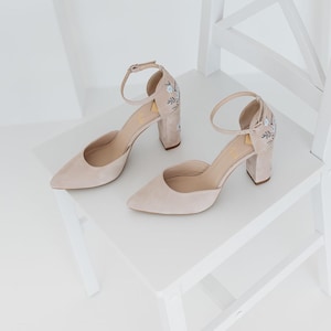 May include: A pair of beige suede block-heel pumps featuring a pointed toe, an adjustable ankle strap with a metal buckle, and delicate floral embroidery in light blue and dark grey thread on the outer side of the heel counter.