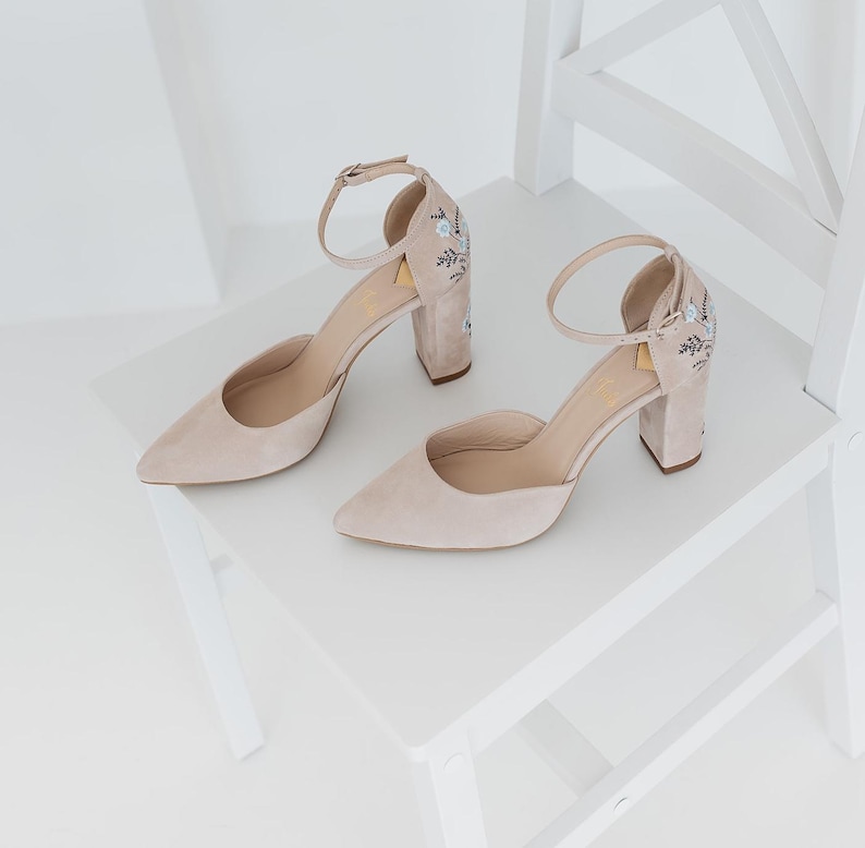 May include: A pair of beige suede block-heel pumps featuring a pointed toe, an adjustable ankle strap with a metal buckle, and delicate floral embroidery in light blue and dark grey thread on the outer side of the heel counter.
