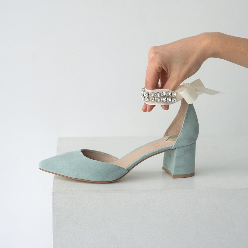 Puede incluir: Un zapato de tac&oacute;n bajo y punta afilada en color azul claro. El calzado cuenta con una tira de cinta blanca en el tobillo adornada con una hilera de cristales transparentes brillantes. El exterior es mate y tiene un dise&ntilde;o lateral abierto.