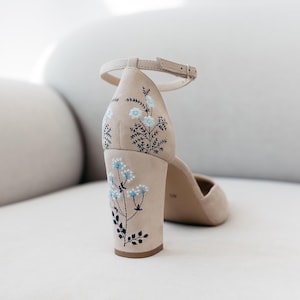 May include: A beige suede block-heel shoe featuring an ankle strap with a metal buckle. The heel and back of the shoe are decorated with delicate embroidery of light blue flowers and dark green stems, extending from the base up to the heel.