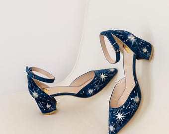 May include: A pair of navy blue ankle strap heels with a starburst pattern. The shoes have a pointed toe, a small block heel, and a buckle closure. The starburst design is in white and gold.