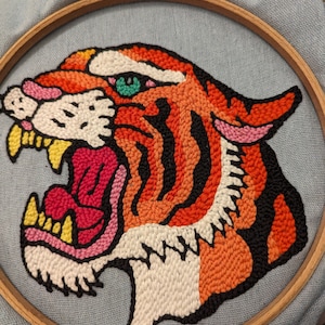 Può includere: Ricamo a mano di una testa di tigre. La tigre presenta dettagli arancioni, neri, bianchi e rosa, con un occhio verde e zanne gialle. Il ricamo è fissato in un cerchio di legno su uno sfondo di tessuto azzurro.