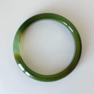Puede incluir: Un brazalete redondo de jade verde. El brazalete pulido tiene una superficie lisa y un aspecto translúcido, con variaciones de tonos verdes y amarillos. La joya está sobre un fondo blanco.