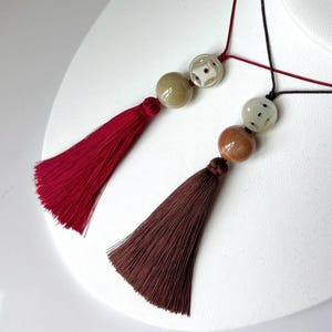 Puede incluir: Dos collares con borlas exhibidos sobre una superficie blanca. Uno presenta una borla burdeos con una cuenta beige y estampada. El otro tiene una borla marr&oacute;n con una cuenta marr&oacute;n y estampada. Ambos tienen cordones negros y rojos.