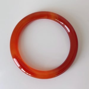 Peut inclure: Un bracelet jonc en agate orange et rouge translucide. Le bracelet circulaire est une bande pleine de pierre polie, présentant un dégradé de couleurs allant de l'orange clair au rouge foncé. Un bijou simple et élégant.