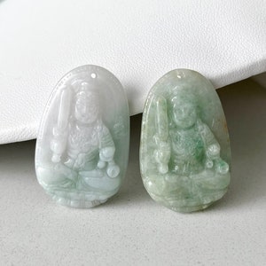 以下が含まれることがあります： それぞれ剣を持った座像が彫られた2つの翡翠ペンダント。ペンダントは楕円形で、1つは淡い緑色、もう1つは緑色の縞模様が入った半透明の白色です。背景は白い表面です。