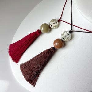 Puede incluir: Dos collares con borlas y cuentas. Uno tiene una borla burdeos y una cuenta verde claro, el otro tiene una borla marr&oacute;n y una cuenta marr&oacute;n. Ambos collares tienen una cuenta blanca con un dise&ntilde;o geom&eacute;trico.